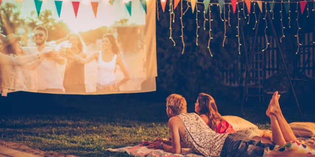 Young couple having movie night party. Laying down on blanket in front of movie improvised screen. Backyard decorated with festive string lights. Night time.