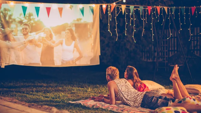 Pareja joven celebrando una noche de cine. Tumbados en una manta delante de una pantalla de cine improvisada. Patio trasero decorado con luces festivas. De noche.