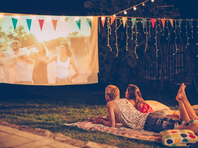 Young couple having movie night party. Laying down on blanket in front of movie improvised screen. Backyard decorated with festive string lights. Night time.