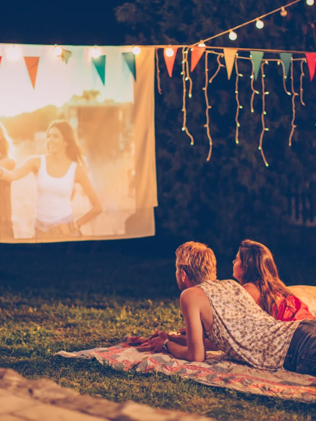 Young couple having movie night party. Laying down on blanket in front of movie improvised screen. Backyard decorated with festive string lights. Night time.